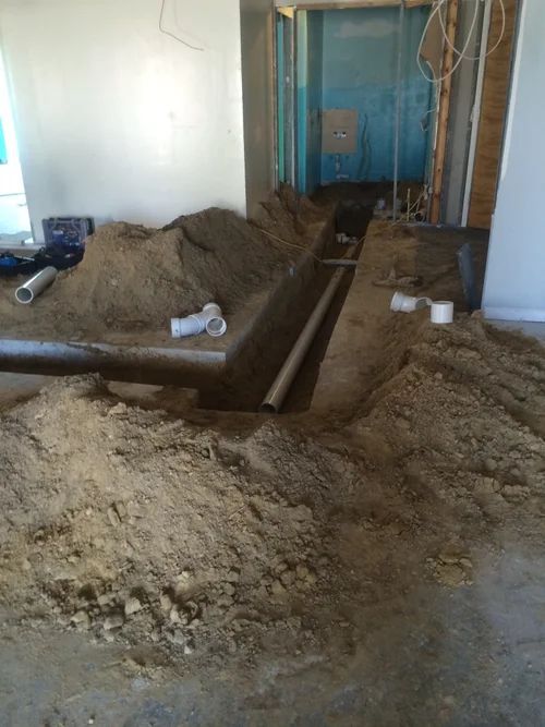 Construction site interior with trench, pipes, and dirt piles. Wall studs, wiring, and white walls are visible.
