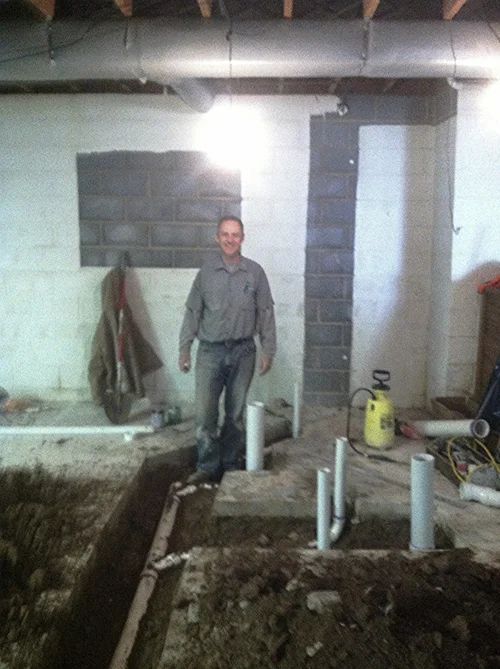 Man stands in a basement, amidst plumbing work. Dirt trench, pipes, and a yellow sprayer are visible.