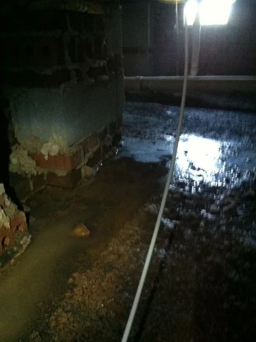 Flooded crawl space with standing water near brick foundation and light source.