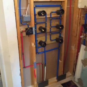 Plumbing rough-in with blue and red PEX pipes, black valves, and electrical box within a wall cavity.