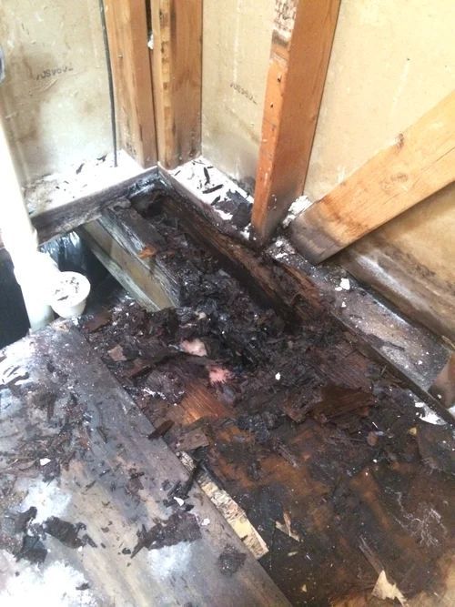 Damaged bathroom floor with rotted wood, showing a dark, wet area around the pipes.