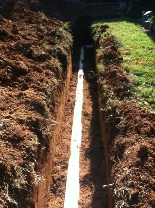 Trench with a white pipe laid inside, surrounded by brown soil and green grass.