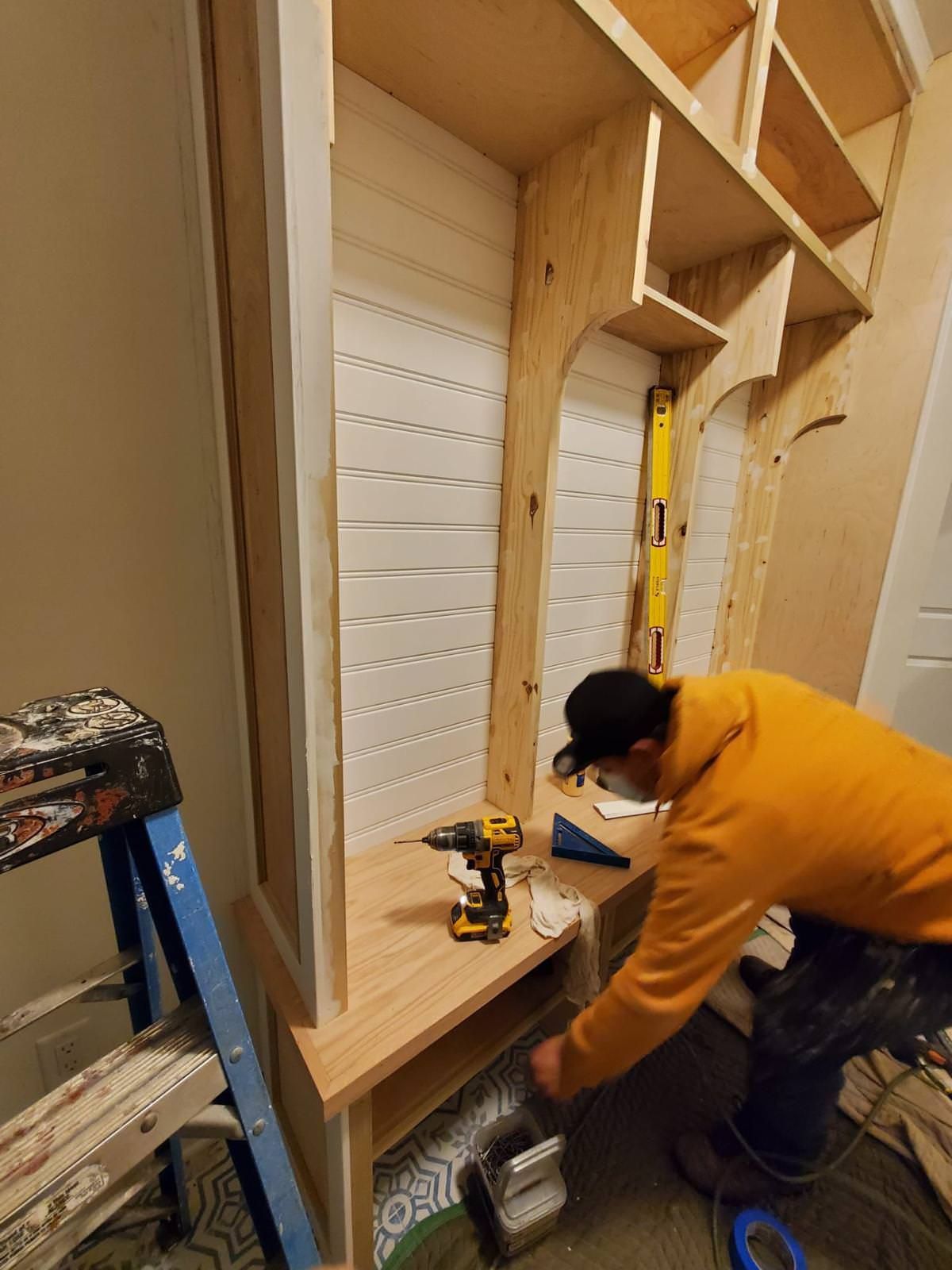 Person assembling a wooden cabinet frame in a workshop, using a drill near a blue ladder and yellow level