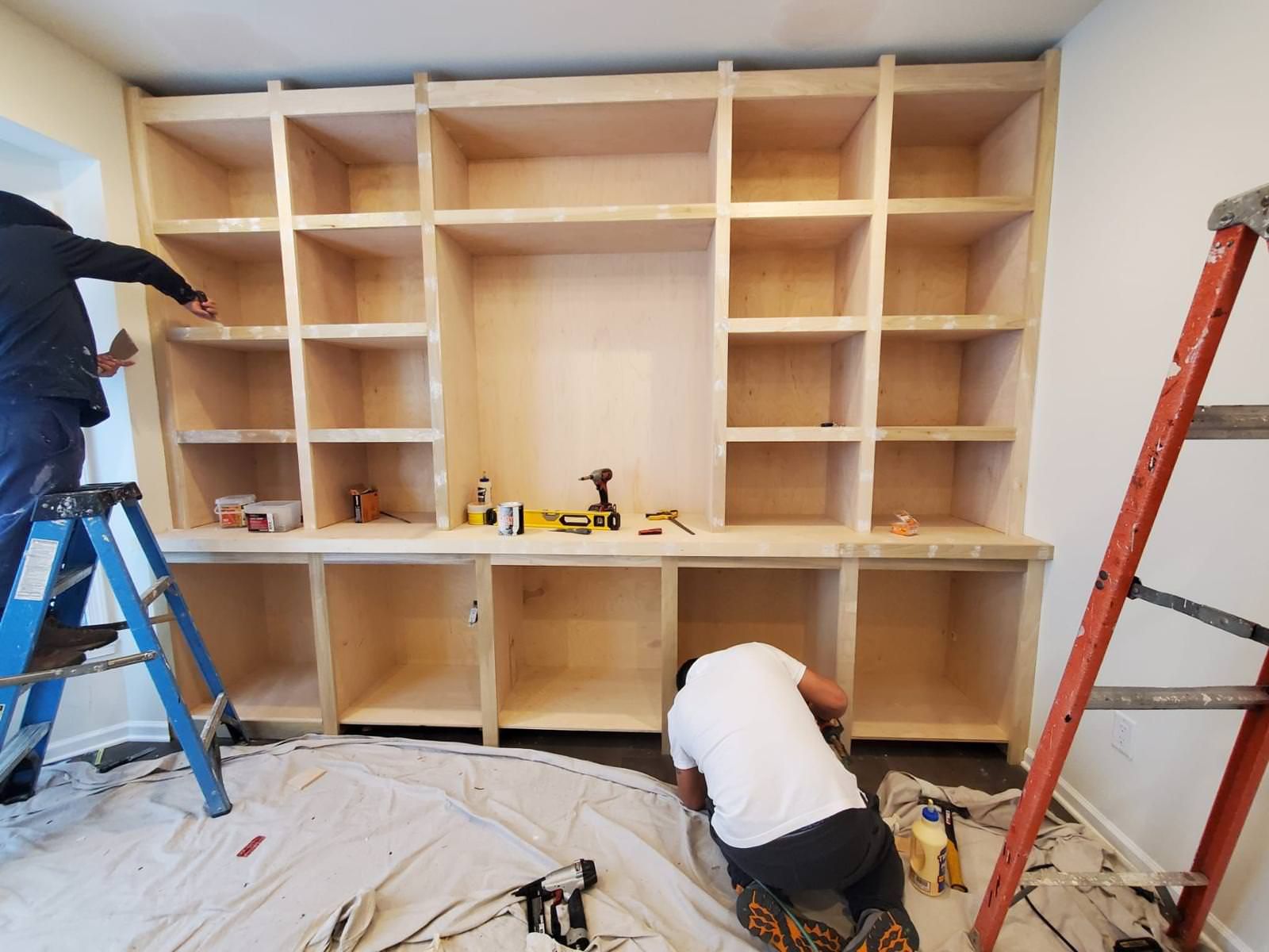 People assembling a large built-in wooden shelving unit with ladders in a room