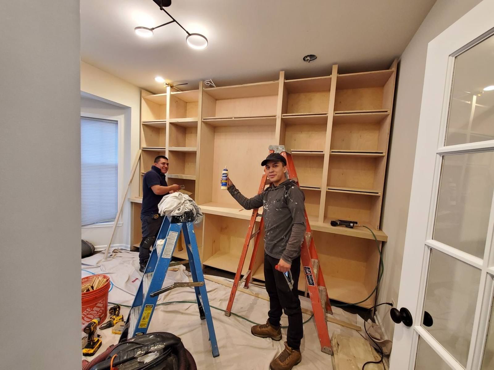Two workers on ladders assembling tall wooden shelving in a room under renovation