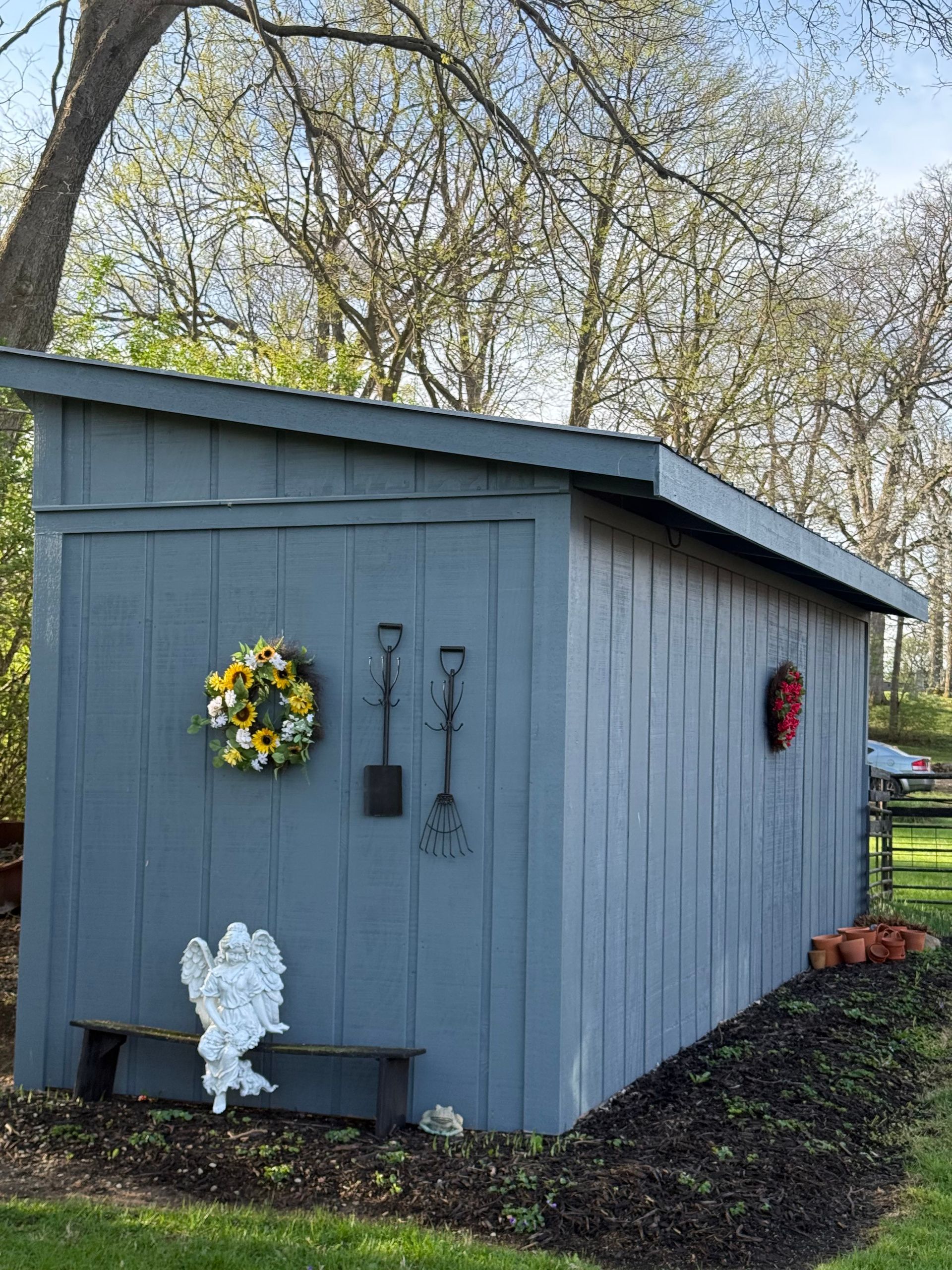 Gray garden shed with decorative wreaths and a white statue in a wooded yard