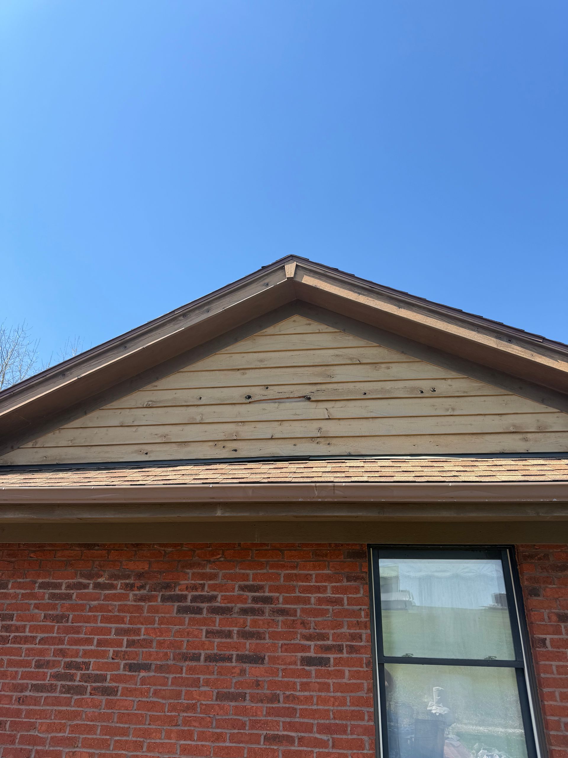 Exterior of a house with a red brick wall, window, and tan gabled roof against a blue sky