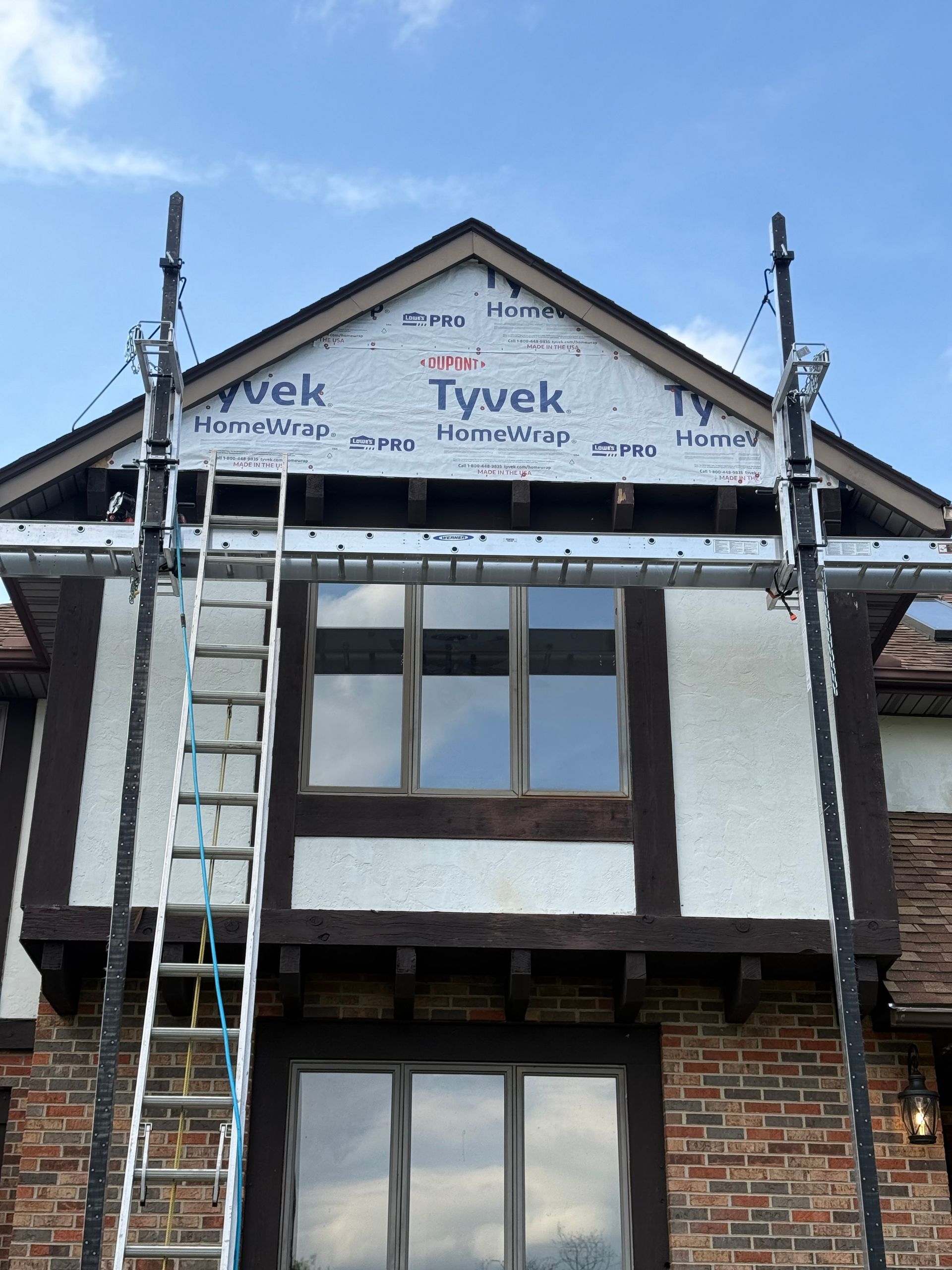 House exterior under renovation with scaffolding, ladder, and Tyvek house wrap on the upper gable.