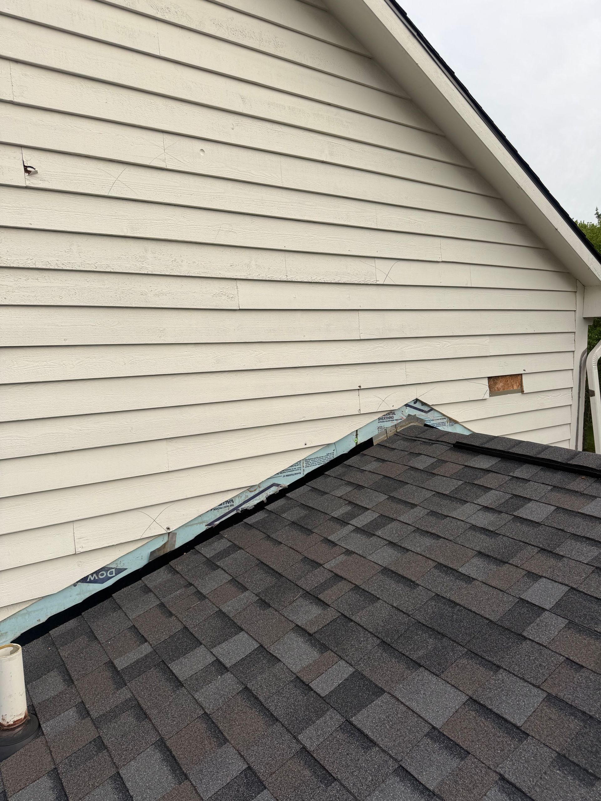 Roof edge beside white house siding with dark gray shingles and blue trim from a close-up angle