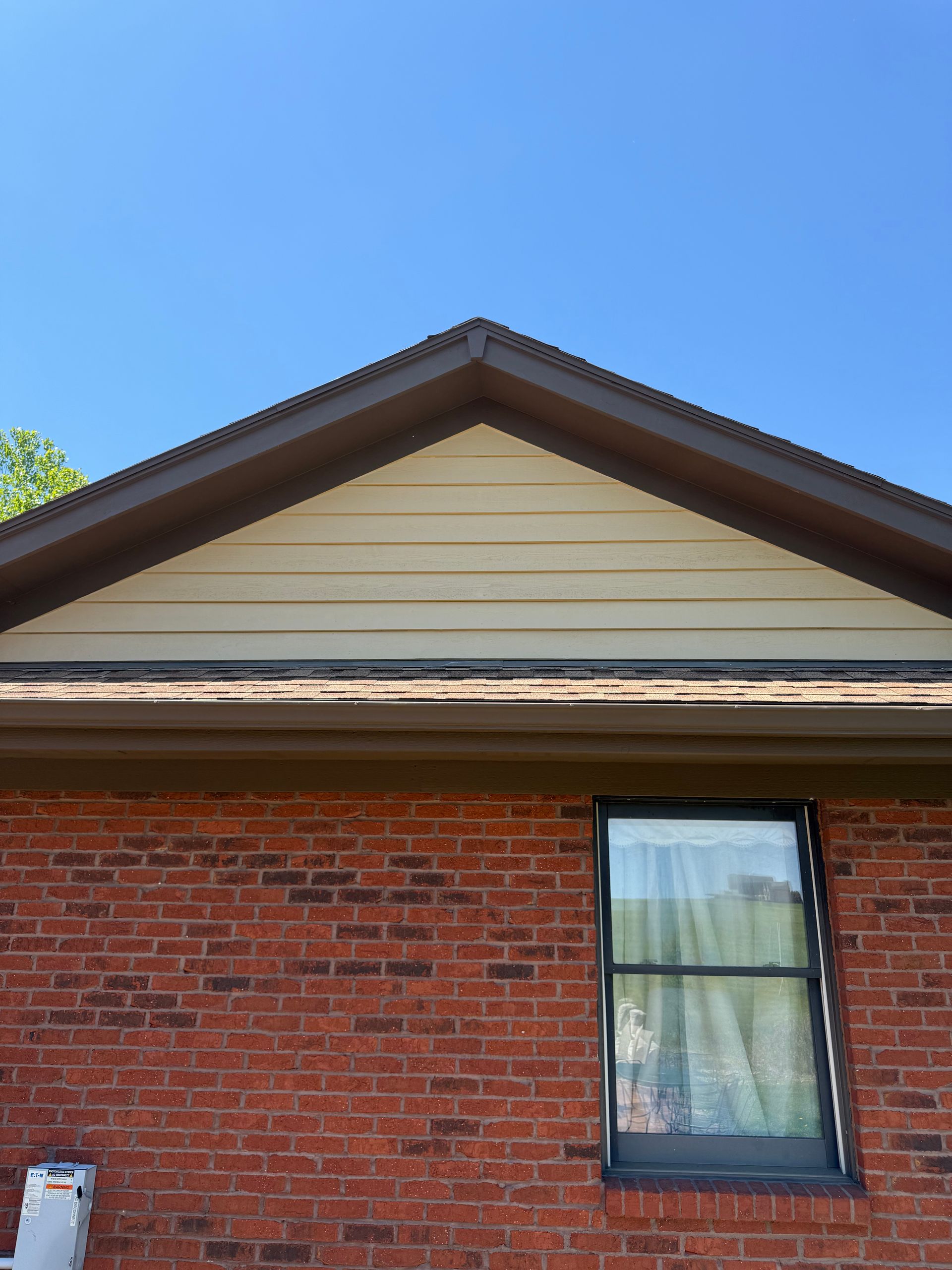 Brick house wall with a single window, tan gable siding, and blue sky above