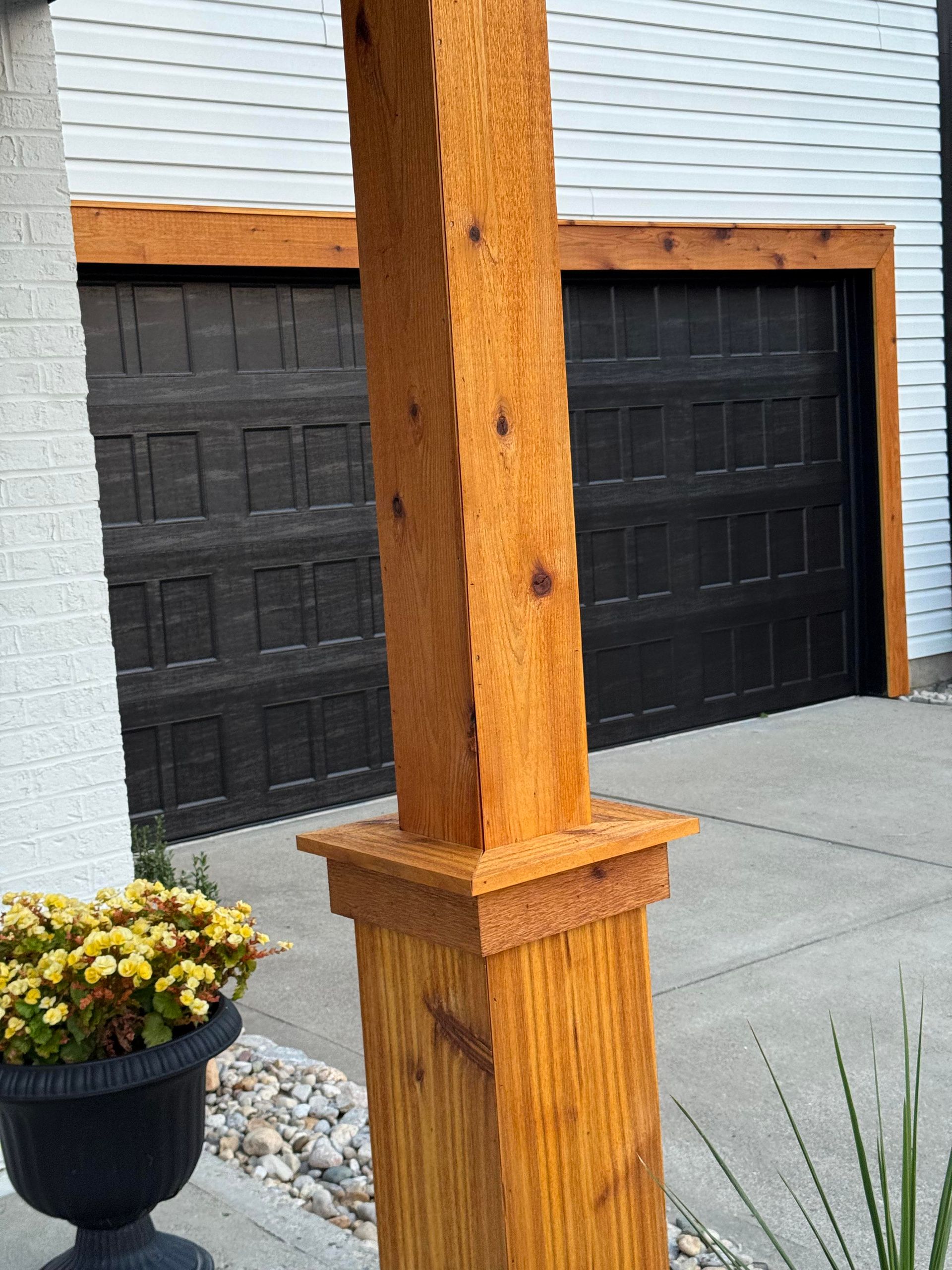 Wooden porch post in front of two dark garage doors and a potted flower on a concrete driveway