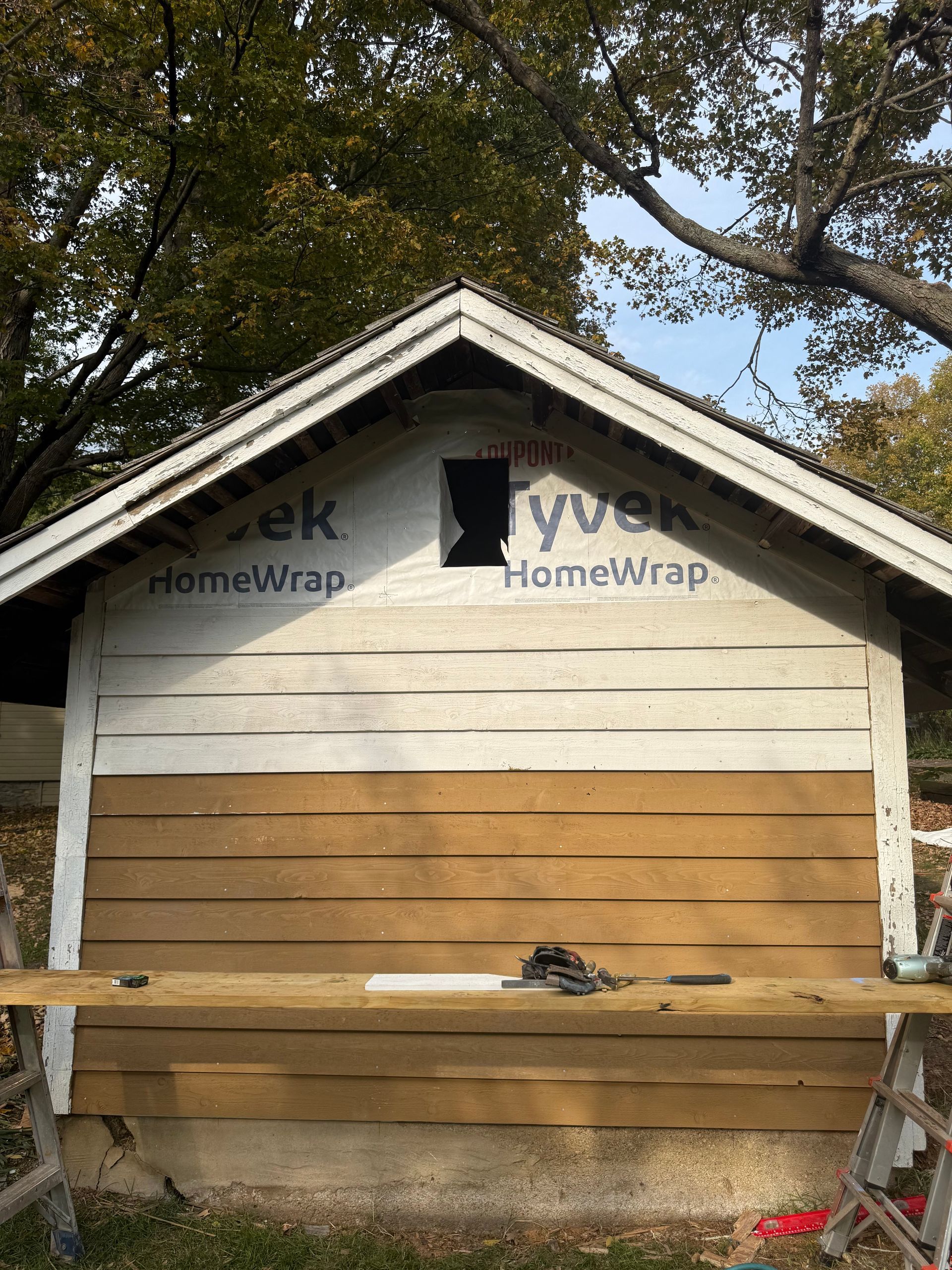 Small tan wooden shed with gabled roof under trees, unfinished siding and a workbench in front