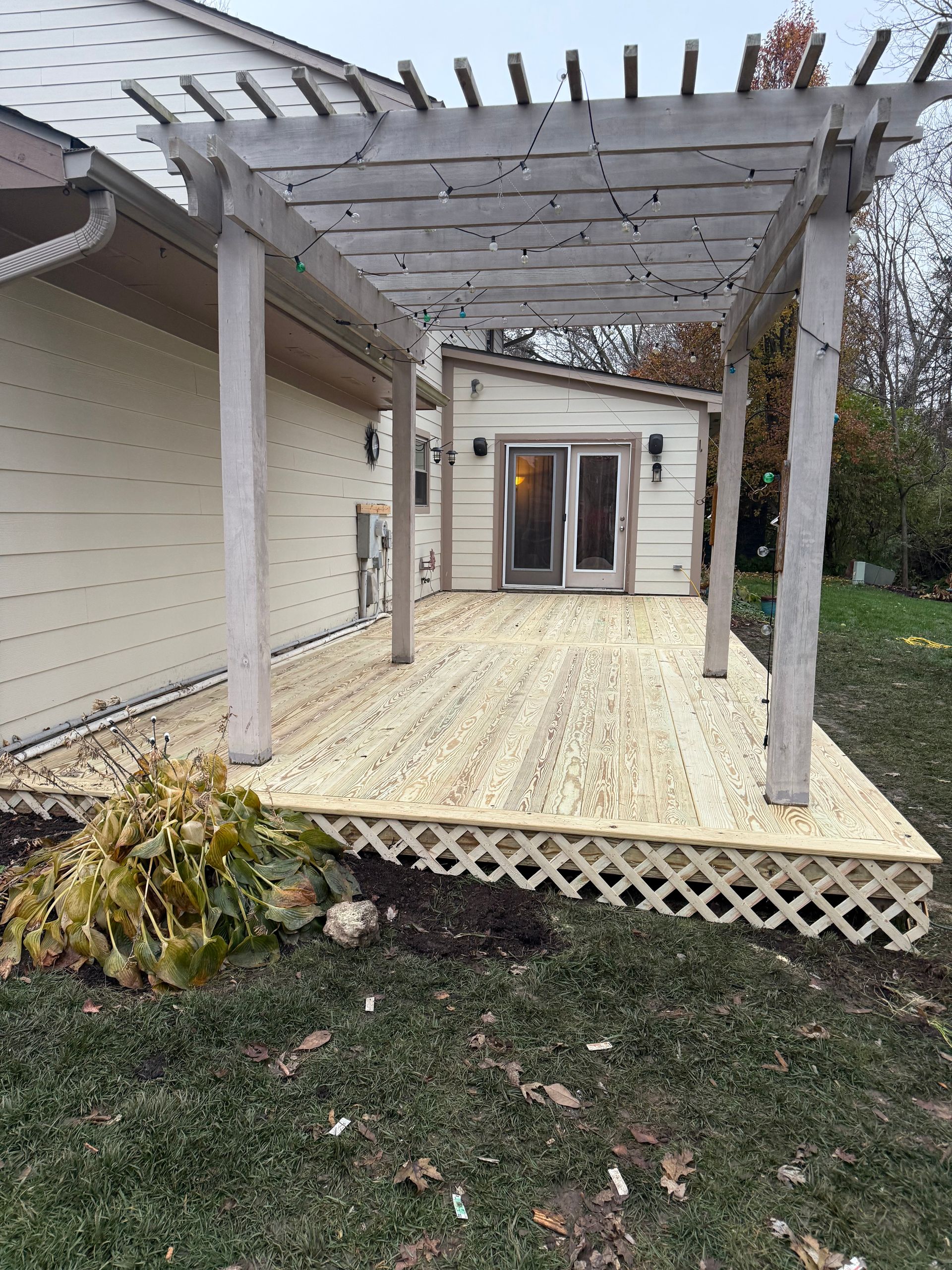 Backyard wooden pergola and deck beside a house, with lattice skirting and patio doors.