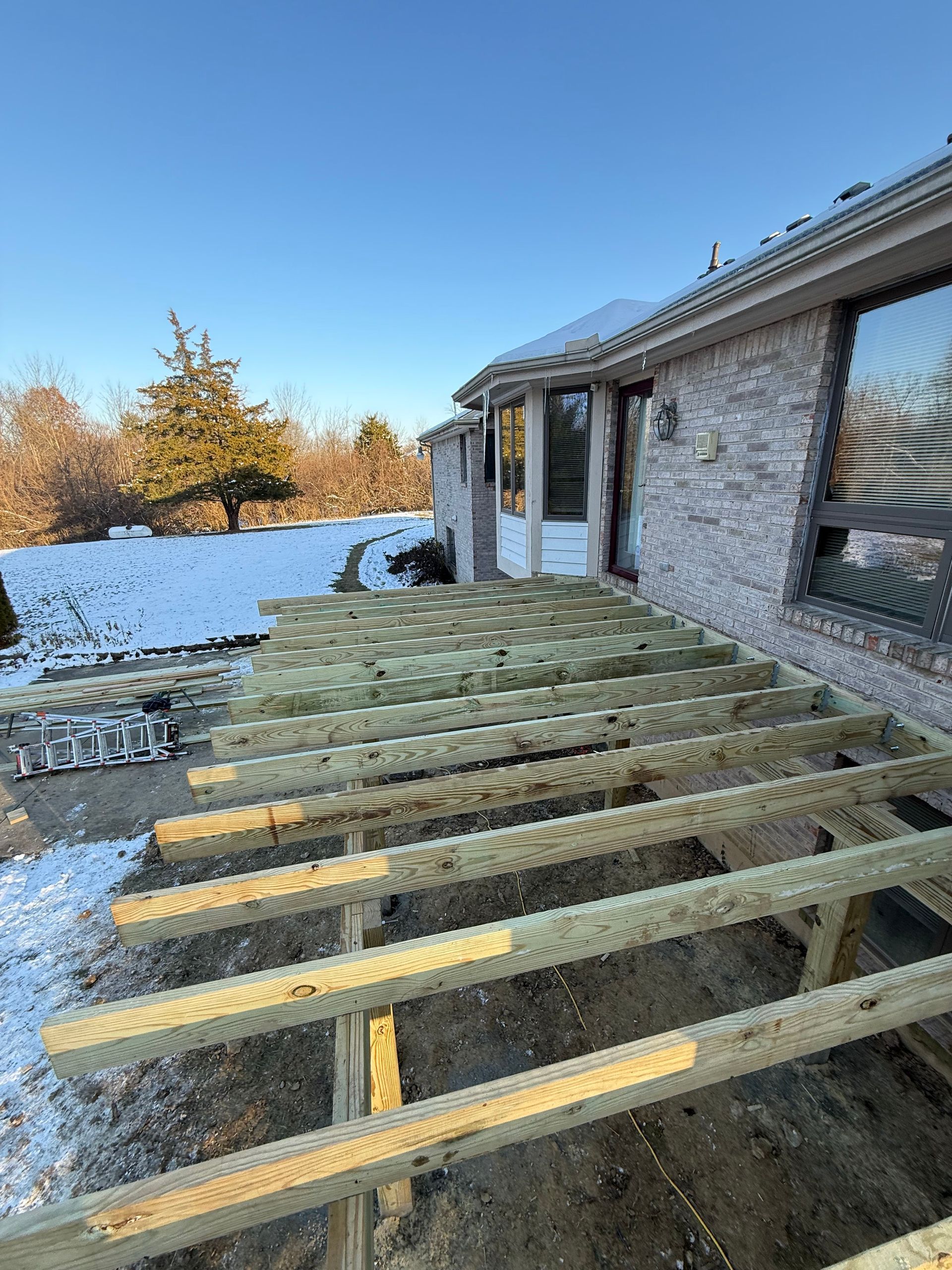 Unfinished wooden deck framing beside a stone house in a snowy backyard at sunset
