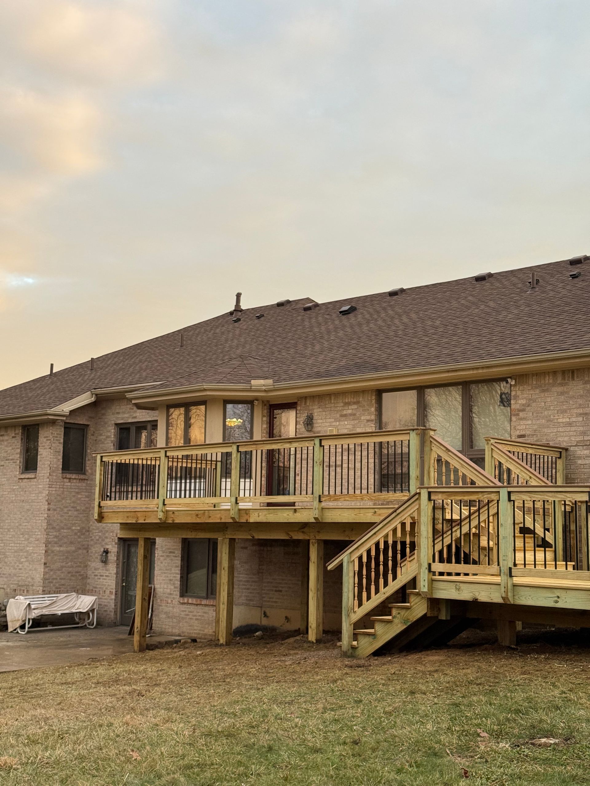 Backyard deck attached to a two-story stone house at sunset, with stairs and a small lawn