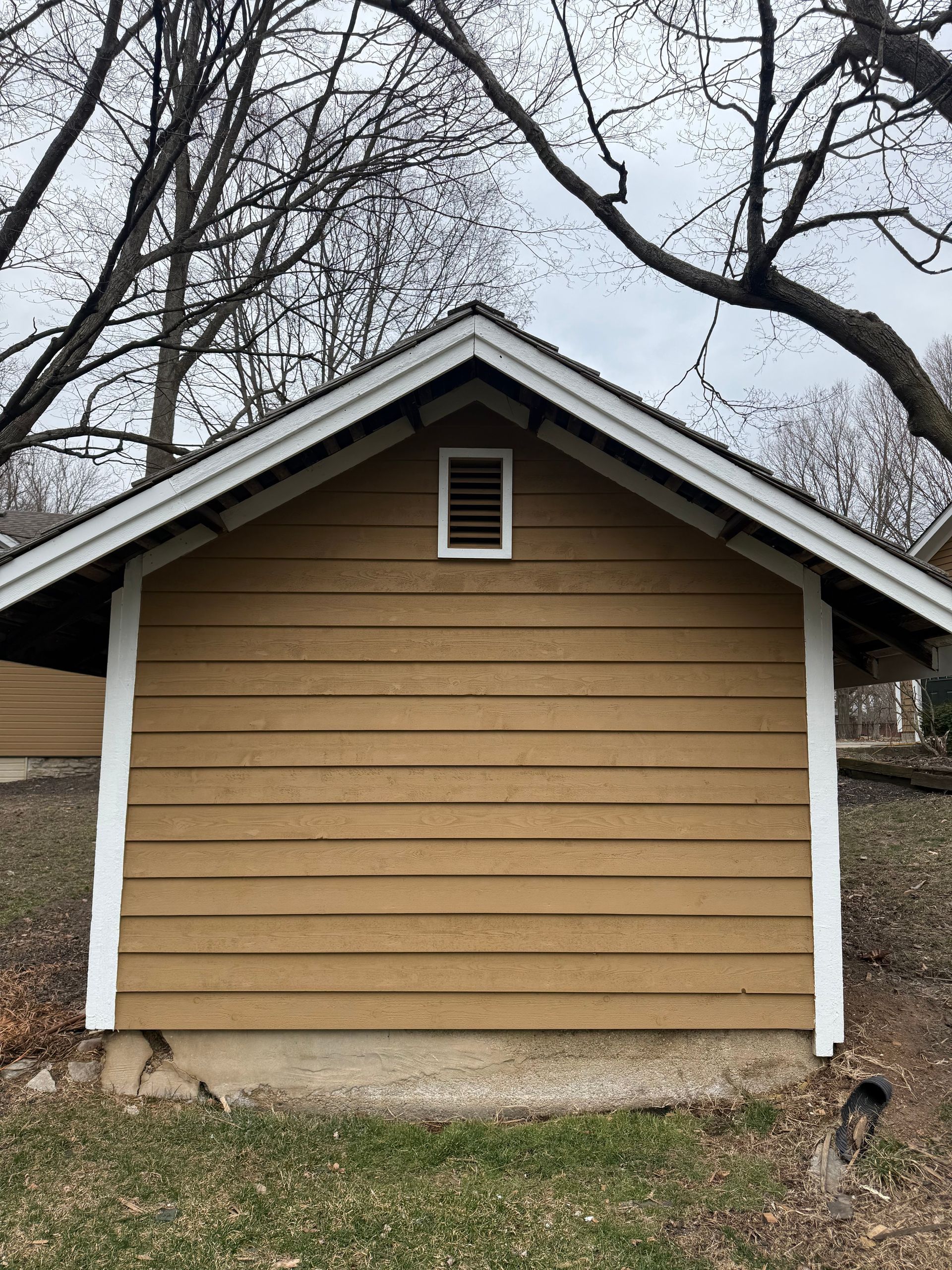 Tan house exterior with white trim and a small vent, framed by leafless tree branches.
