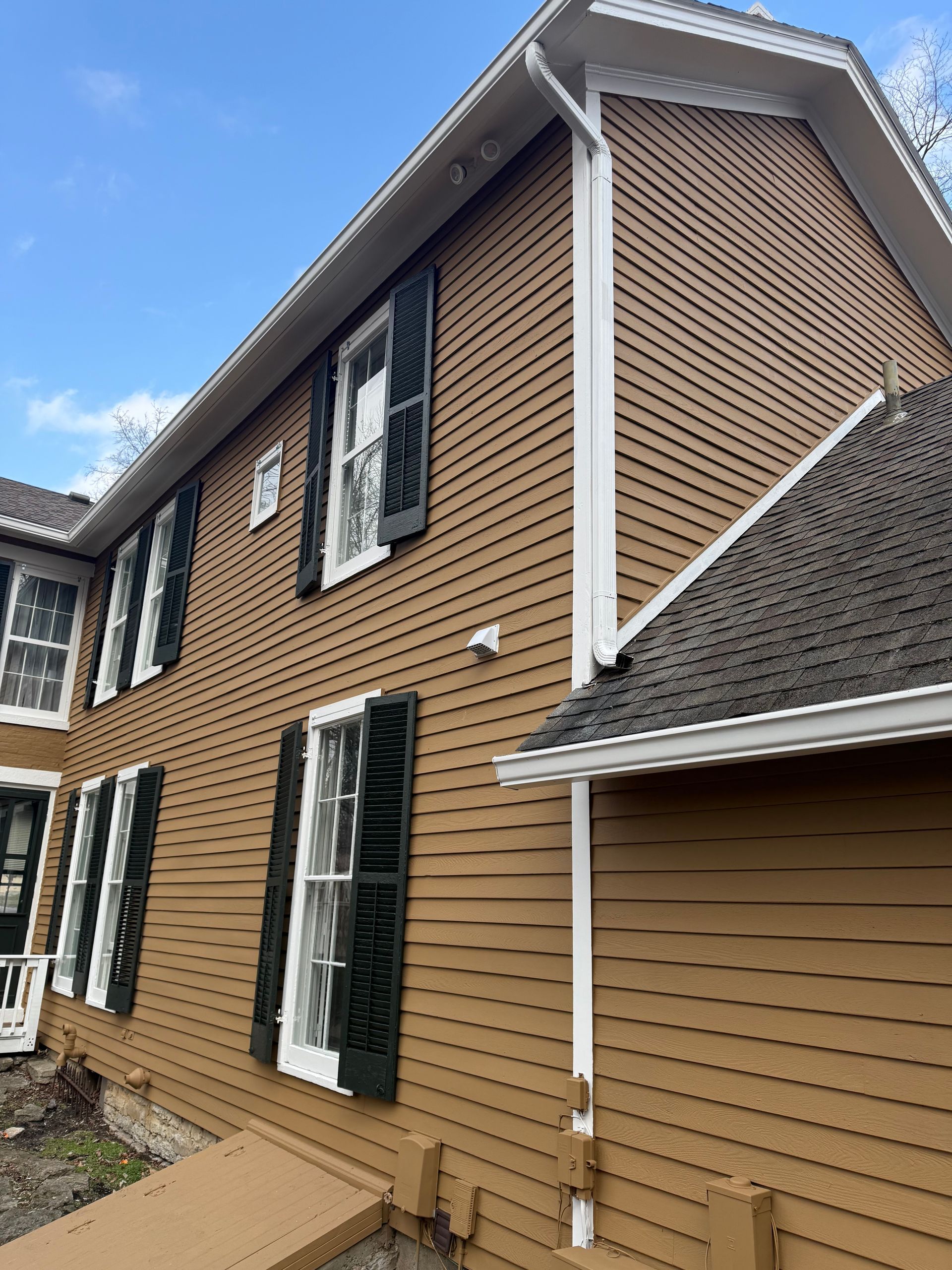 Two-story tan house with white trim, black shutters, and brown shingle roof under a blue sky