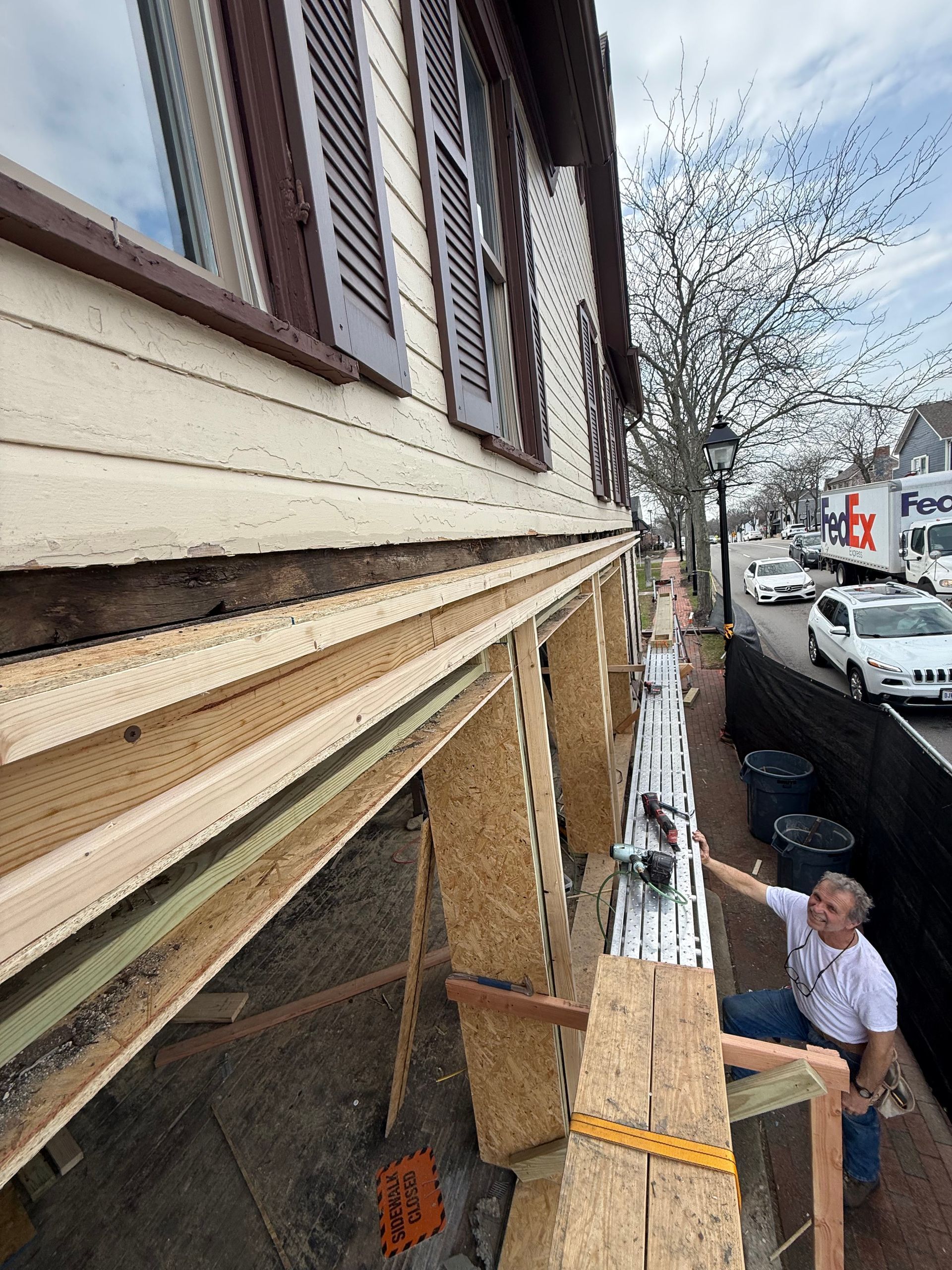 Workers installing wood siding on a house exterior beside a street, with scaffolding and materials stacked below