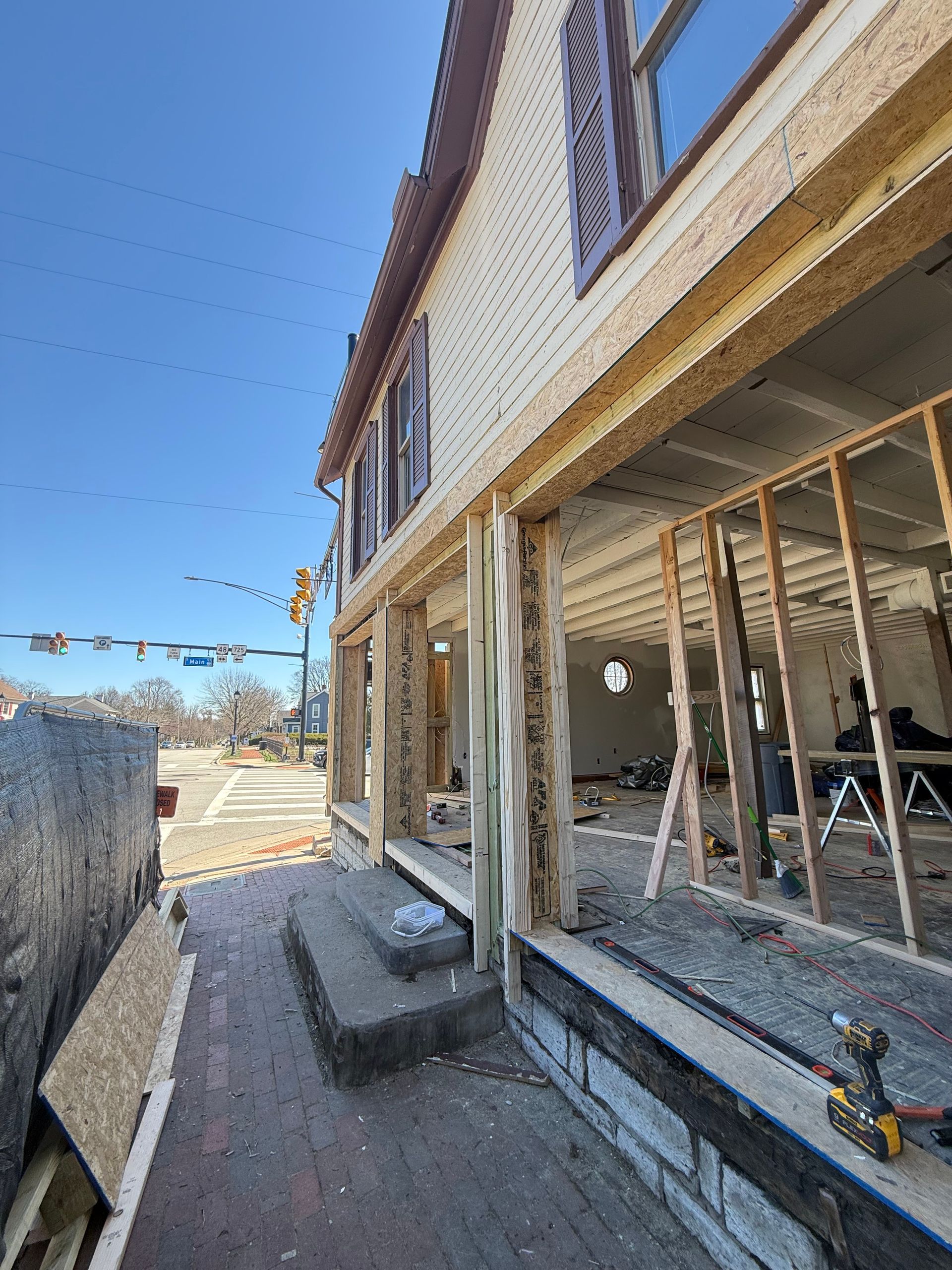 Exterior of a house under construction with exposed framing, stone facade, and workers’ tools on a narrow walkway.