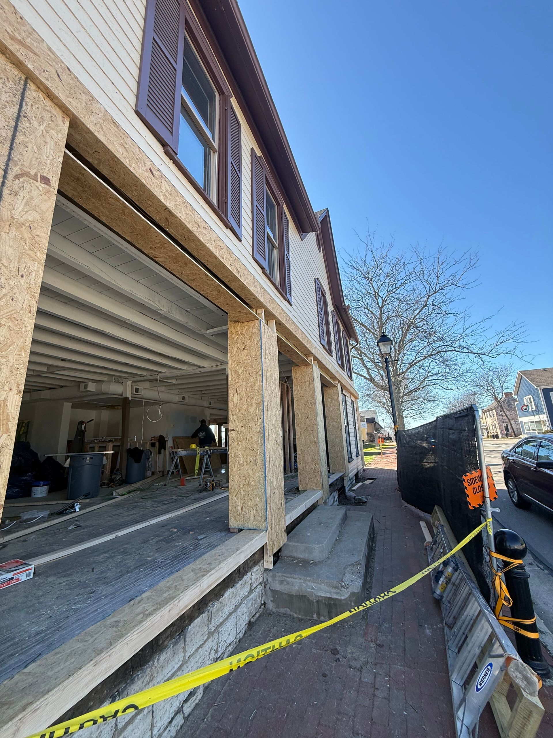Damaged building facade beside a sidewalk, with debris, caution tape, and a clear blue sky.