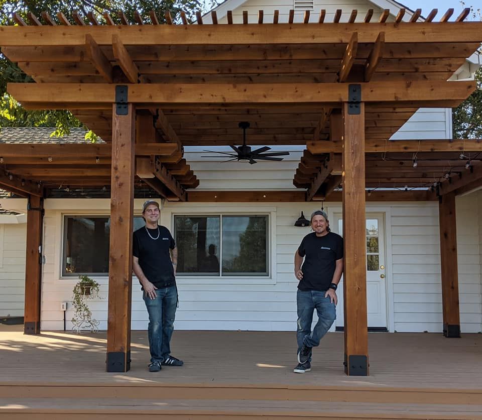 Two men standing under a new wooden pergola on a deck outside a white house.