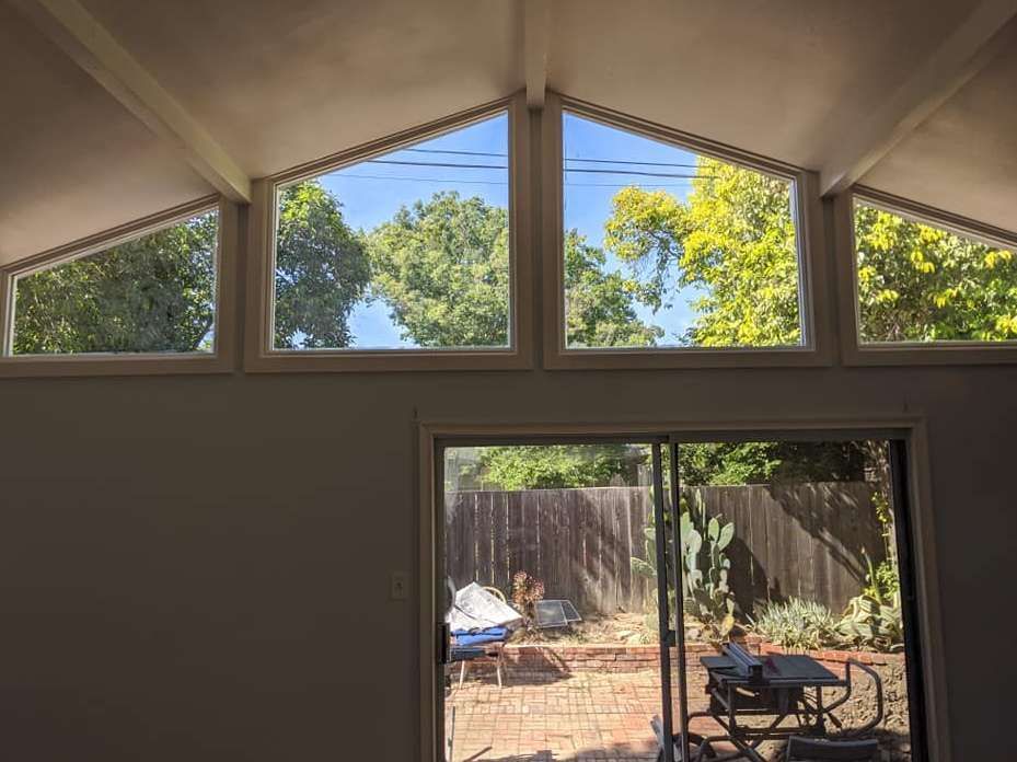 Interior view with large triangular windows and sliding glass door, looking onto a backyard with trees.