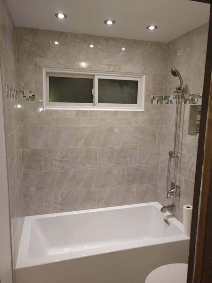 Bathroom with white tub, grey tile walls, showerhead, window, and recessed lighting.