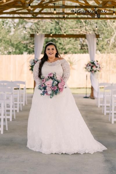 Bride in a white lace gown holding a pink bouquet at an outdoor wedding altar