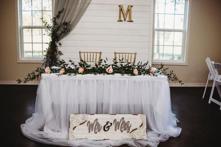 Wedding sweetheart table with white tulle skirt, floral garland, and gold monogram under windows
