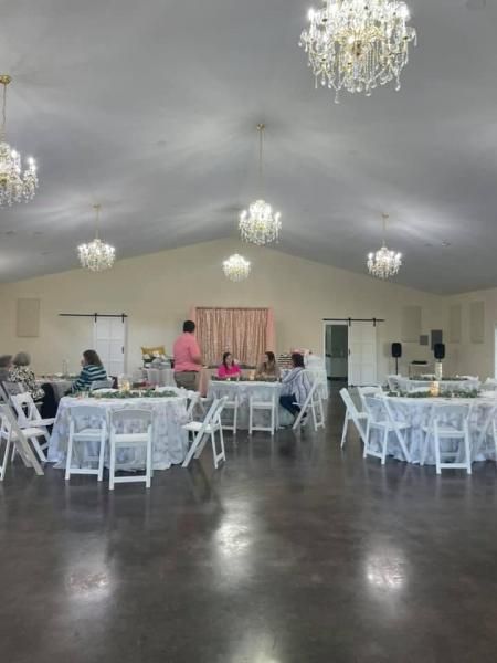 Elegant banquet hall with white tables and chairs, chandeliers, and a few people setting up for an event