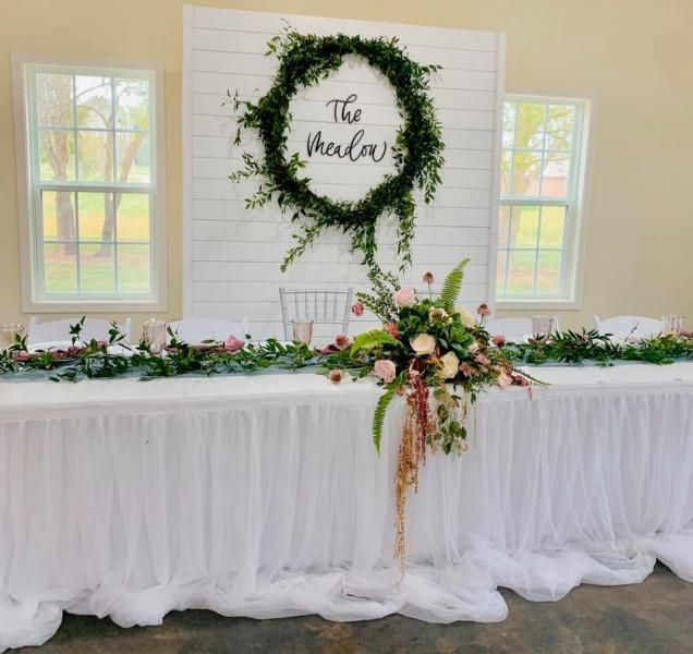 Decorated wedding reception table with white draping, greenery, floral arrangement, and wreath sign backdrop