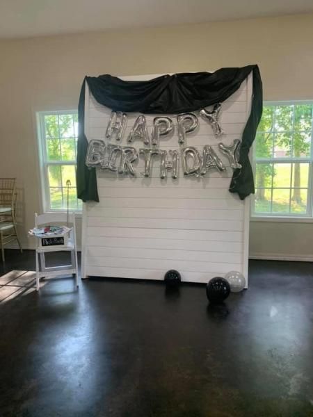 Birthday backdrop with silver “Happy Birthday” balloons, black drapes, and white shiplap wall in a room