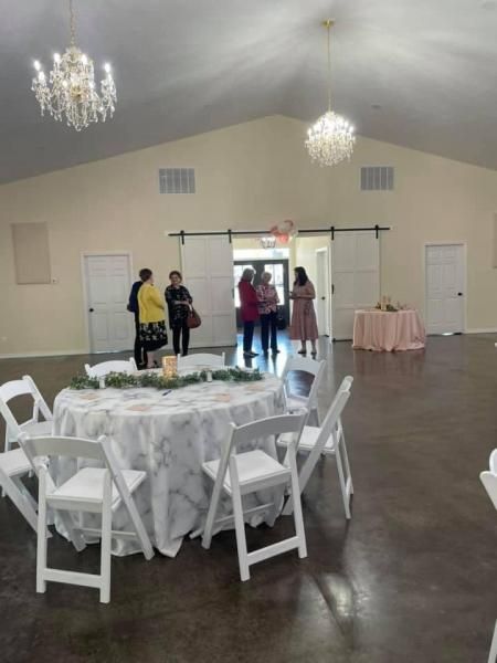 Decorated banquet hall with round tables, white chairs, chandeliers, and a few people near the back wall.
