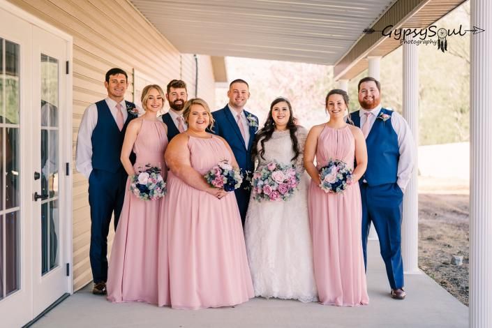 Wedding party posing under a porch, with bridesmaids in pink dresses and groomsmen in blue suits