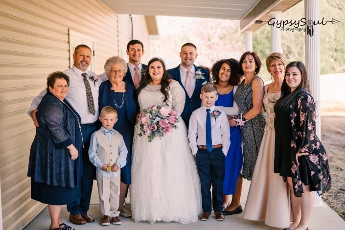 Wedding group posing on a porch, with the bride holding a bouquet surrounded by family and guests.