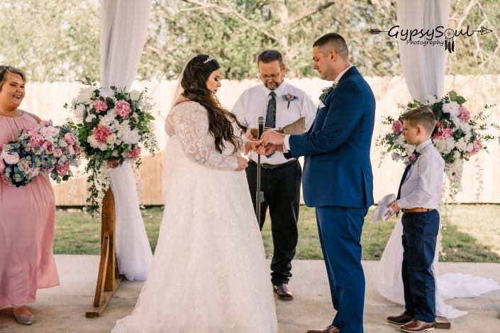 Wedding ceremony with couple exchanging rings under a decorated outdoor canopy, flanked by floral arrangements.