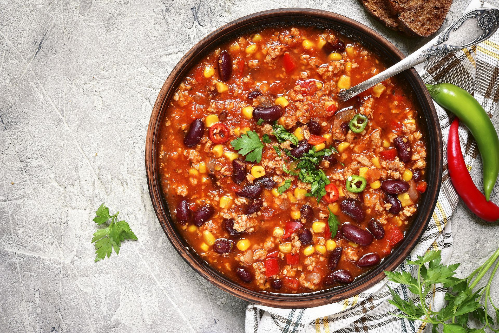A top-down view of a bowl of chili with red beans, corn, and ground meat, garnished with herbs, next to chili peppers.
