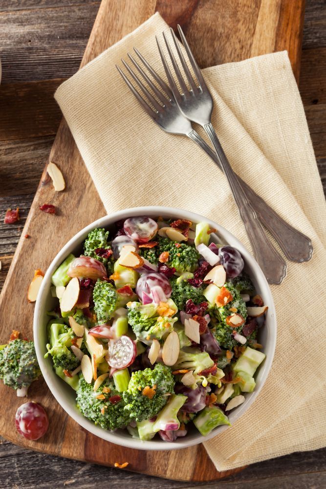 A bowl of broccoli and grape salad with slivered almonds, served on a napkin with two forks on a wooden surface.