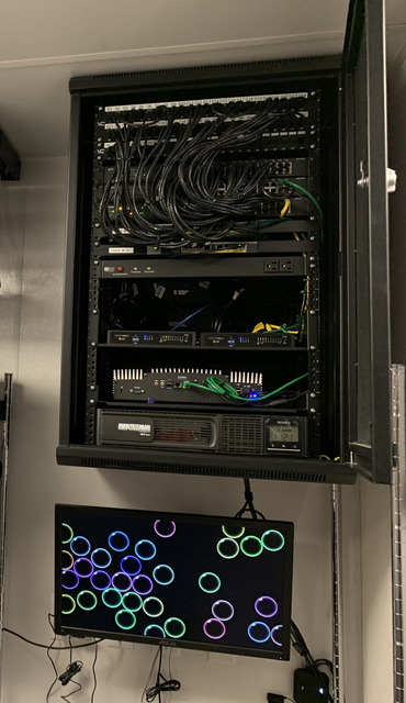 Wall-mounted server rack containing electronic equipment and wires above a monitor displaying colorful circles.