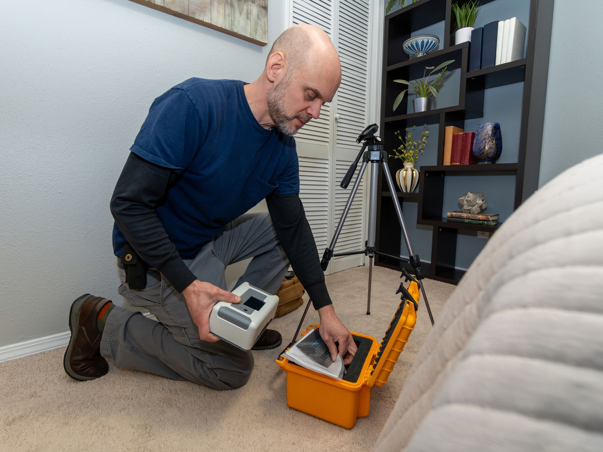 Man kneels, opening a yellow case, with a device in hand, indoors, near a bed and bookshelf.