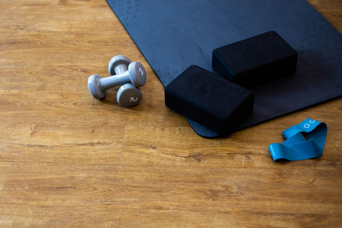 Yoga mat with black foam blocks, silver dumbbells, and a blue resistance clip on a wooden floor
