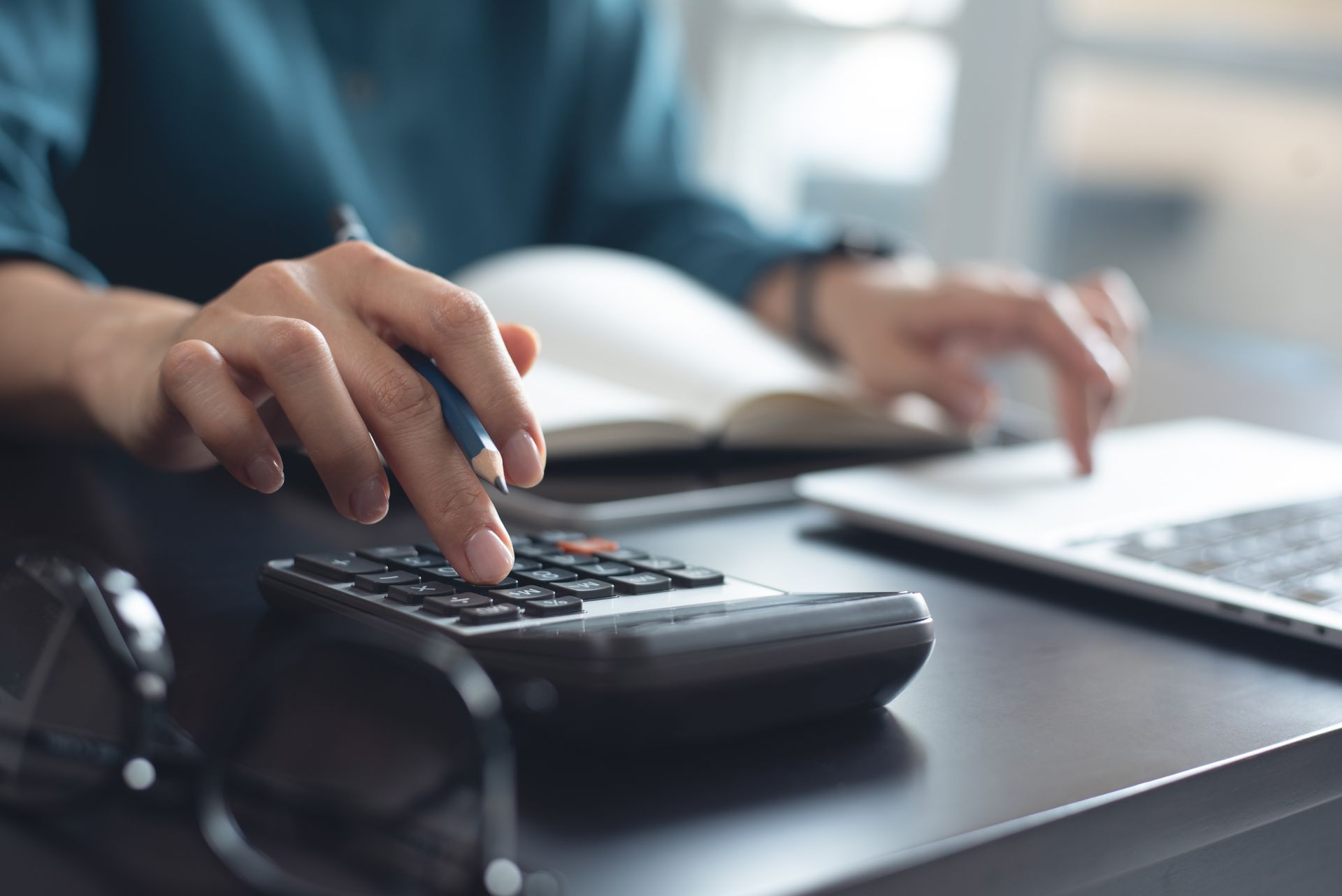 Hand using a calculator beside a laptop on a desk, with an open book in the background Hand using a calculator beside a laptop on a desk, with an open book in the background