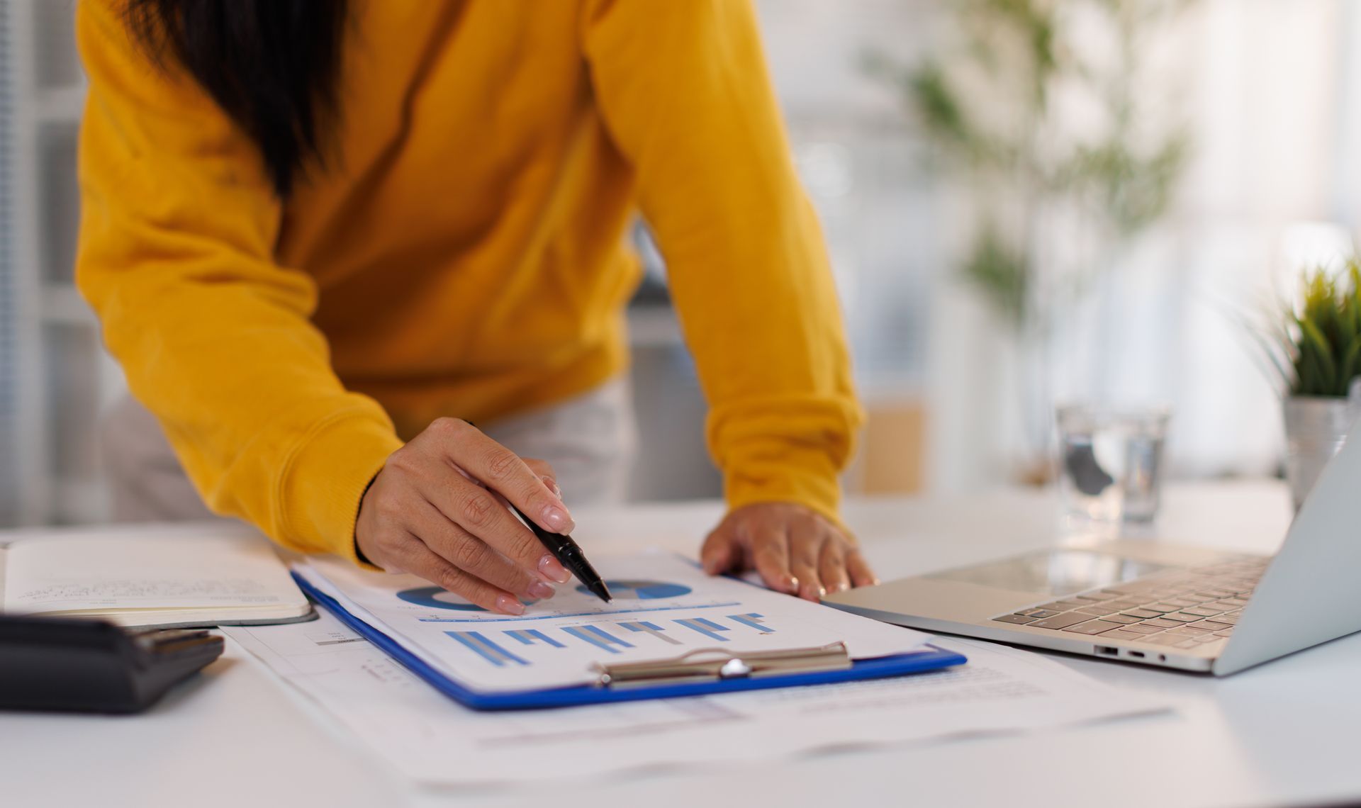 Person in a mustard sweater points to charts on a clipboard beside a laptop at a desk