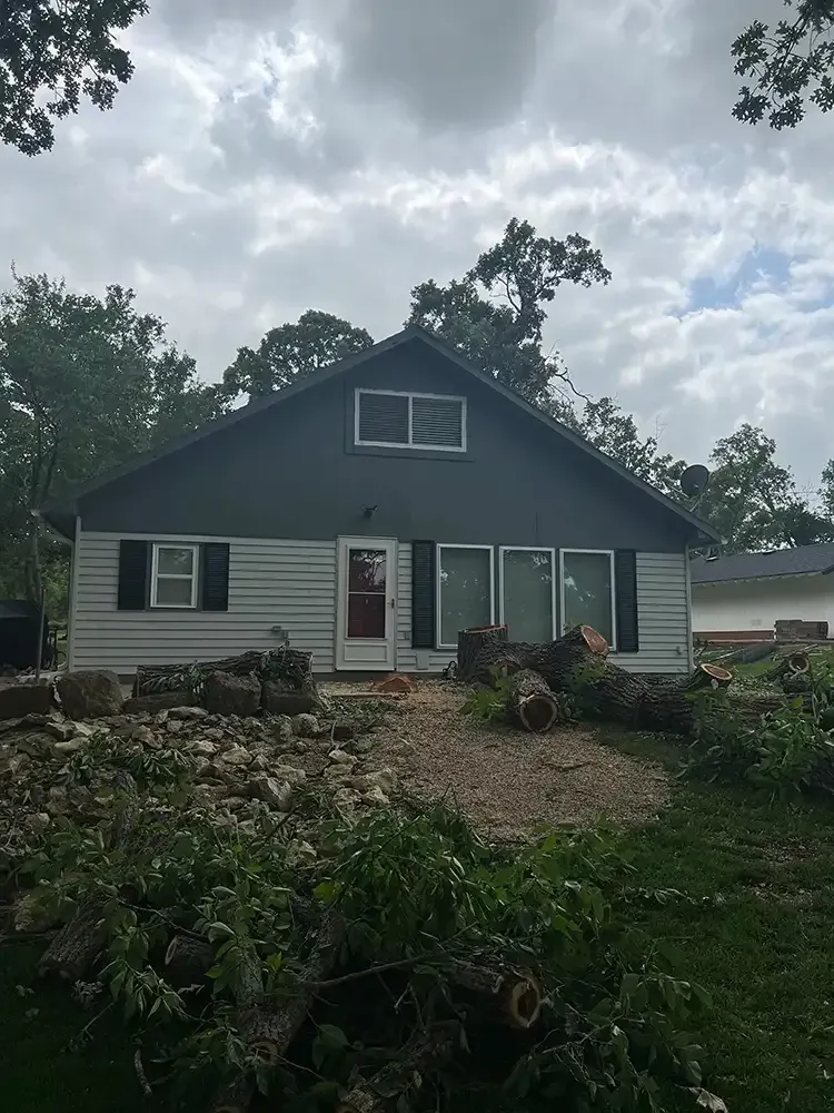 Small white house with dark gray gable roof, green shutters, and storm debris in the yard under cloudy skies