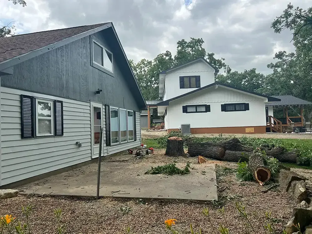 Two gray houses and a white building around a yard with logs and orange flowers under cloudy skies