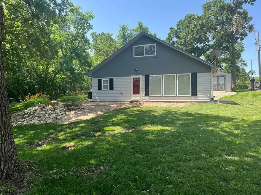 Gray house with red door and large front lawn, surrounded by trees in a sunny yard.