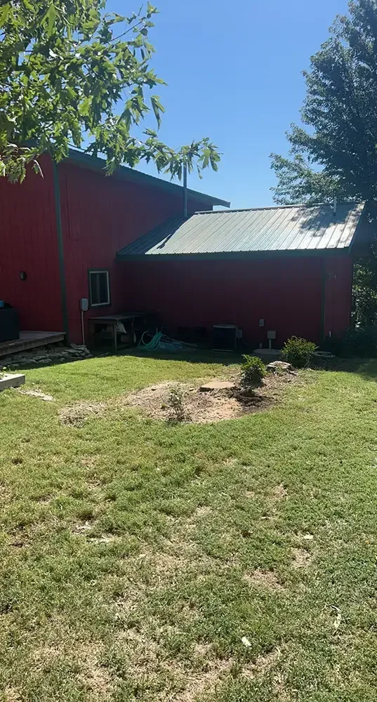 Red barn with gray roof beside a grassy yard and small tree under a clear blue sky