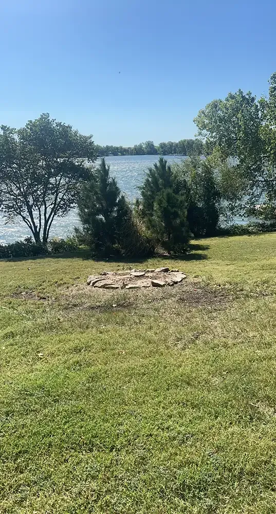 Grassy lakeside yard with trees, a stone fire pit, and water in the background