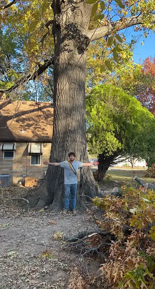 Person standing with arms outstretched beside a large tree in a yard, with a house and autumn foliage behind.