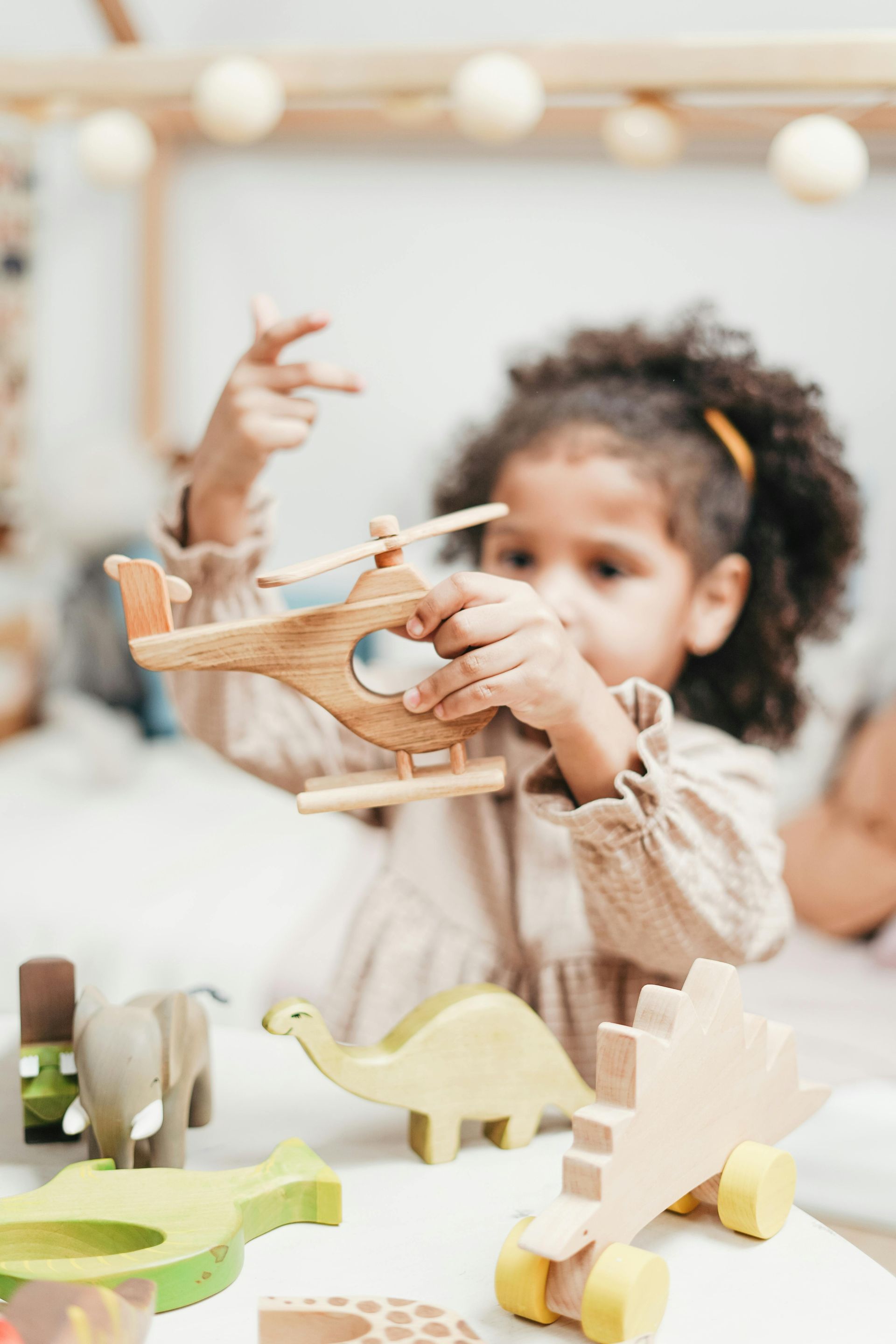 Young girl playing with wooden toys, including a helicopter and dinosaur, in a bright room.